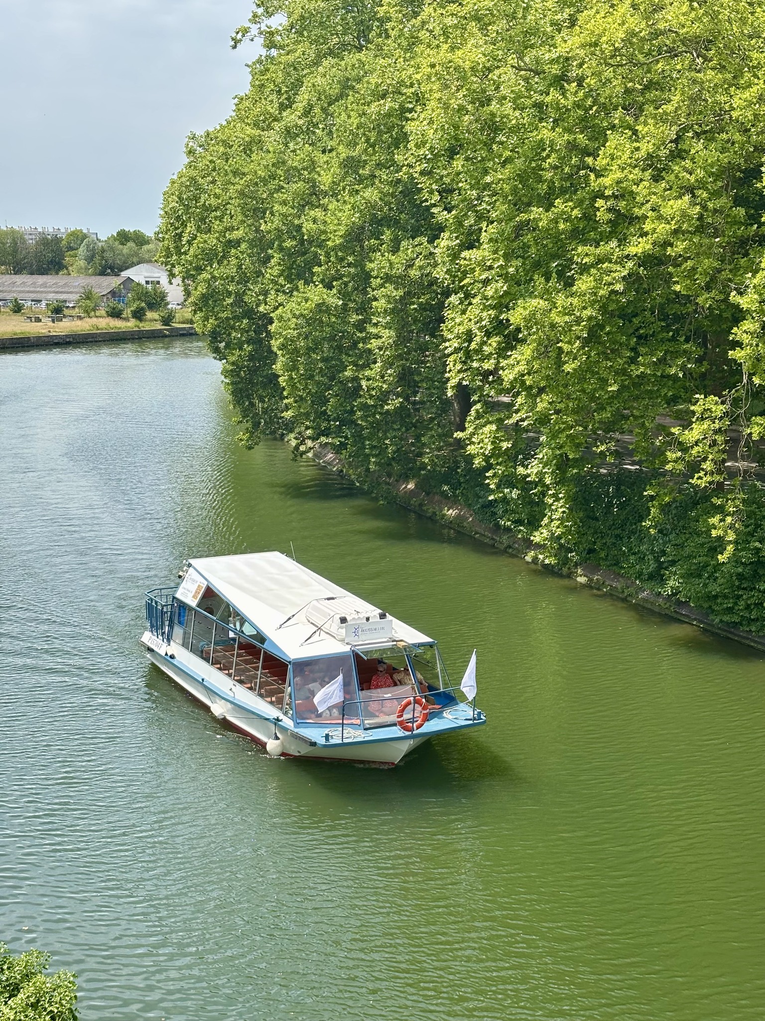 Le bateau Le Moussaillon en navigation sur la Deûle à Lille, le long des berges arborées.